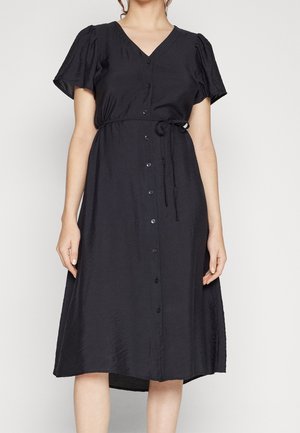 Kvinde iført en sort midi-kjole med korte ærmer, knapper foran, V-hals og et bindebælte i taljen, stående mod en ensfarvet baggrund.