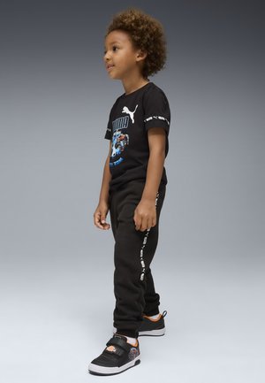 T-shirt noir avec impression graphique, associé à un pantalon de survêtement noir avec des rayures blanches sur les côtés, et des baskets noires avec des accents orange.