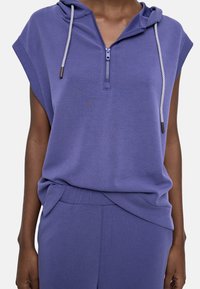 Osoba w fioletowej bezrękawnikowej bluzie z zamkiem z przodu i sznurkami, w zestawie z dopasowanymi fioletowymi spodniami.