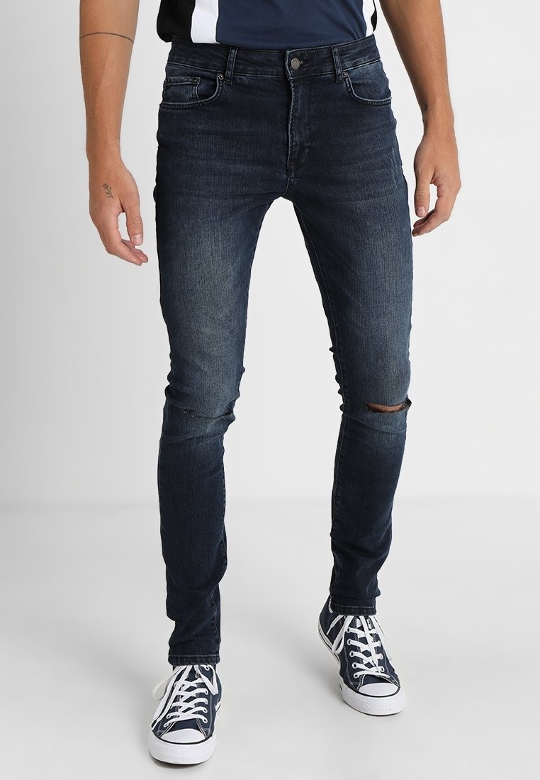 Hombre que lleva jeans oscuros ajustados de color azul con un pequeño rasguño en la rodilla y zapatillas altas de color azul marino con cordones blancos.