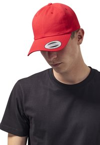 Rode baseballpet met een gebogen klep. De stof is glad met stiksels en een verstelbare band. Gedragen met een simpele zwarte T-shirt.