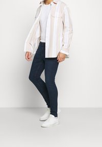 Camisa de manga larga blanca y a rayas pastel sobre una camiseta blanca sencilla, combinada con jeans ajustados oscuros y zapatillas blancas.