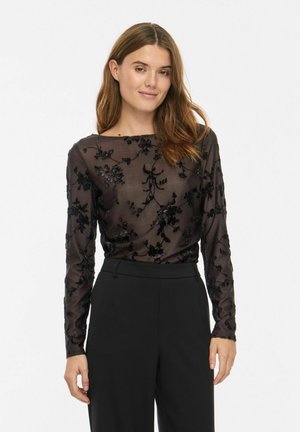 Une femme porte un haut noir transparent à manches longues orné de broderies florales et un pantalon noir, sur un fond blanc uni.