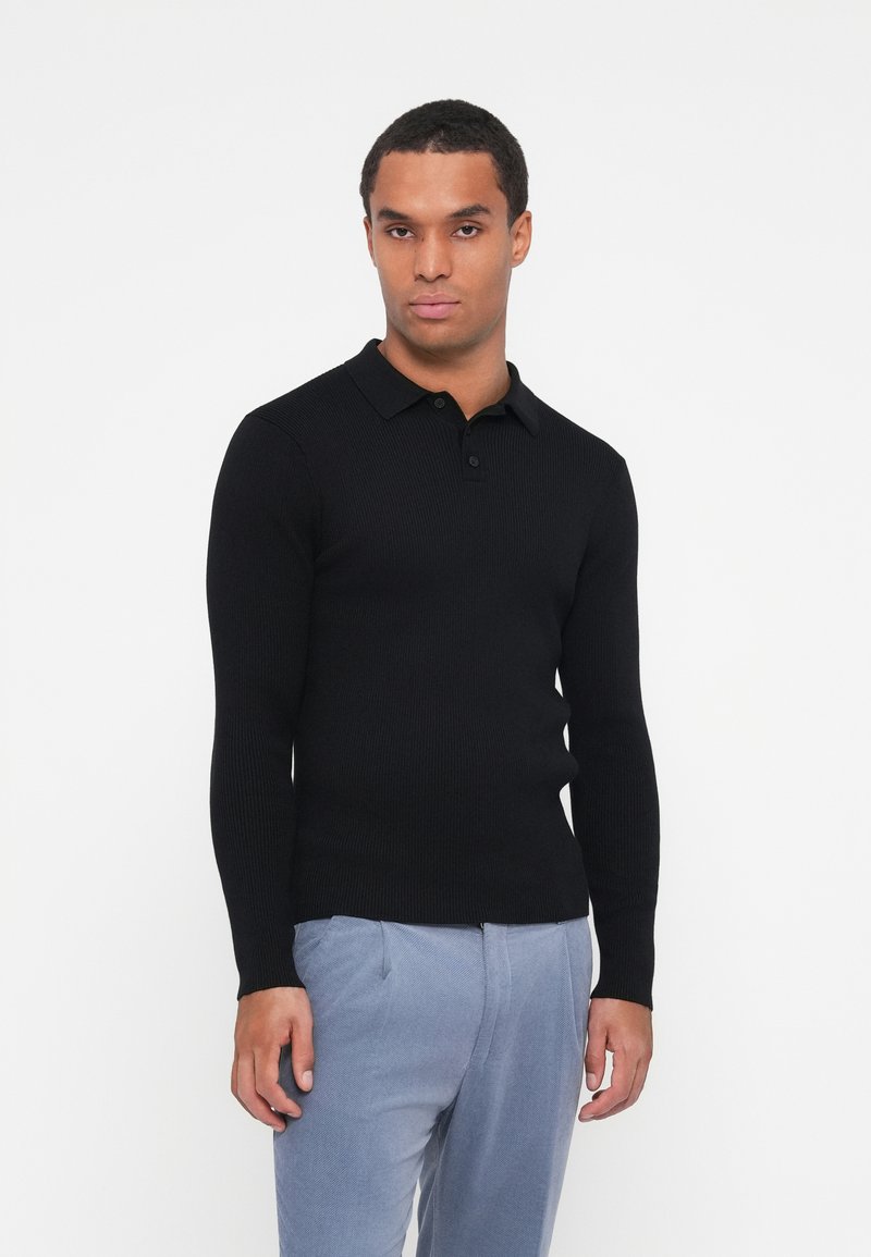 Schwarz geripptes Langarm-Polo-Shirt mit klassischem Kragen und Knopfleiste mit drei Knöpfen, kombiniert mit hellgrauen Hosen.