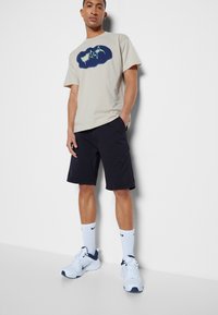 T-shirt bege claro com um gráfico azul escuro, combinado com calções pretos. Meias brancas e sapatilhas brancas com detalhes azuis completam o visual.