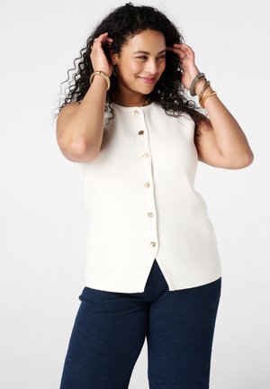 Femme aux cheveux bouclés portant un haut blanc sans manches à boutons et un pantalon bleu marine, souriante et s'ajustant les cheveux avec les deux mains.