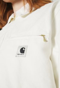 Vit zip-up jacka med en strukturerad yta, som har en liten svart Carhartt-logotyp och en ficka med dragkedja på bröstet med ett guldfärgat drag.