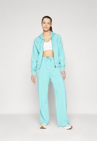 Ανοιχτό μπλε φούτερ με κουκούλα και φερμουάρ μπροστά, σε συνδυασμό με αντίστοιχες χαλαρές παντελόνες. Μαλακό ύφασμα με άνετη εφαρμογή και λεπτομέρεια με κορδόνι.