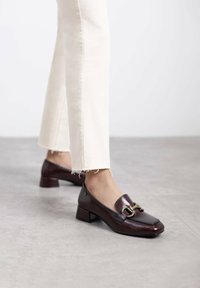 Mocasines de charol marrón con puntera cuadrada y pequeño tacón de bloque, que cuentan con un detalle decorativo de hebilla de metal en el frente.