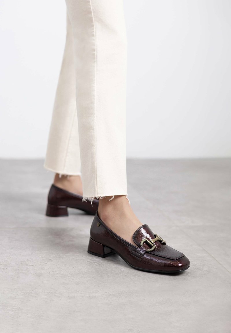 Mocasines de charol marrón con puntera cuadrada y pequeño tacón de bloque, que cuentan con un detalle decorativo de hebilla de metal en el frente.