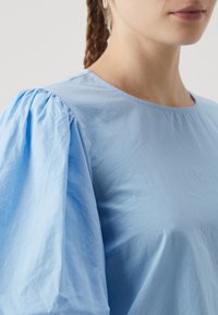 Jasnoniebieska bluzka z bawełny o gładkiej fakturze, z bufiastymi rękawami i okrągłym dekoltem. Bez widocznych wzorów.