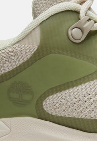 Gros plan sur le tissu beige et vert olive d'une chaussure de sport avec une texture tricotée, un verrou de lacet en plastique et un logo d'arbre en relief sur le panneau latéral.