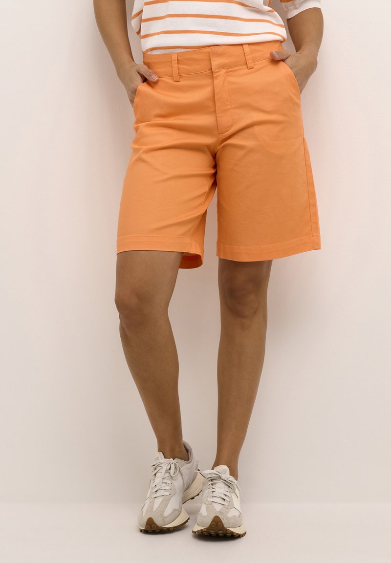 Bermudas de algodón naranja con corte recto, pasadores para cinturón y bolsillos laterales. Llevadas con zapatillas blancas y una camisa a rayas.