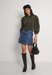 Alyvuogių žalia palaidinė su sagomis ir pūstomis rankovėmis, derinama su džinsine suknele. Juodi aukšti iki kelių batai ir maža juoda rankinė papildo šį įvaizdį.