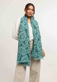 Donna che indossa un lungo sciarpone teal con grandi fantasie floreali, sopra un maglione bianco e pantaloni beige a gamba larga, in piedi davanti a uno sfondo neutro.