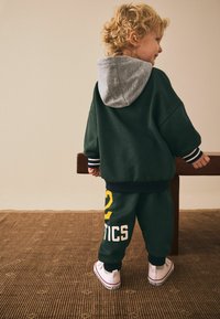 Green hoodie with gray hood, striped cuffs, and matching green sweatpants featuring yellow and white text. White sneakers complete the outfit.