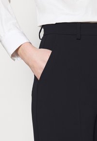 Zwarte dresspants met een perfect aansluitende pasvorm, een gladde textuur en zijzakken. Een witte button-up shirt is gedeeltelijk zichtbaar bij de manchet.