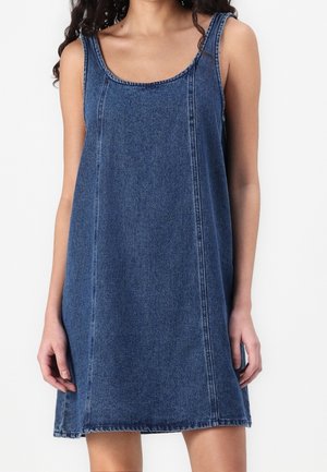 Woman wearing sleeveless blue denim dress with scoop neckline and vertical seams, standing against white background.