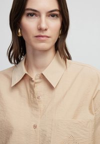 Beige button-up shirt with a textured fabric, featuring a pointed collar and a decorative pocket detail with eyelet stitching.