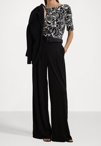 Woman wearing black wide-leg pants, black woven belt, black and white floral top, black blazer draped over shoulder, with hands in pockets.