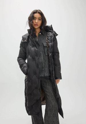 Mujer con un abrigo largo negro acolchado sobre un suéter de punto gris oscuro y pantalones anchos a juego, de pie sobre un fondo claro y liso.