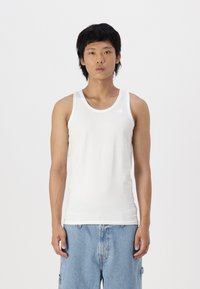 Hvit bomullstopp med rund hals og tynne stropper, kombinert med løse blå denimshorts, stående foran en ensfarget bakgrunn.