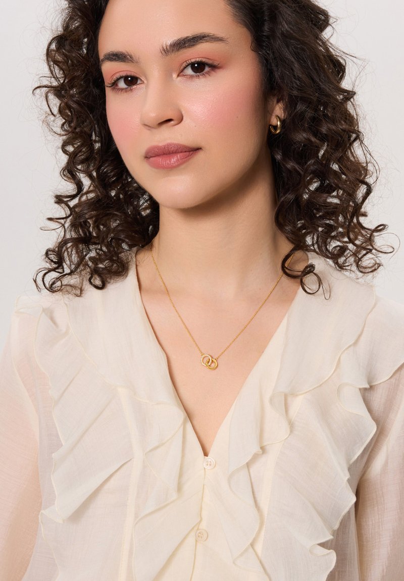 Złoty naszyjnik z przeplatanymi okręgami, noszony na bluzce z kremowego materiału z falbankami i dekoltem w kształcie litery V.