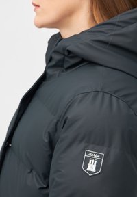 Zwarte pufferjack met een capuchon, voorzien van een logopatch op de linker schouder en een gladde, gewatteerde textuur.