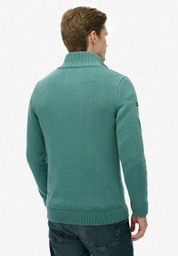 Turkusowy sweter z dzianiny z wysokim kołnierzem, prążkowanymi mankietami i dołem. Materiał wydaje się miękki i teksturowany, o dopasowanym kroju i minimalnych zdobieniach.