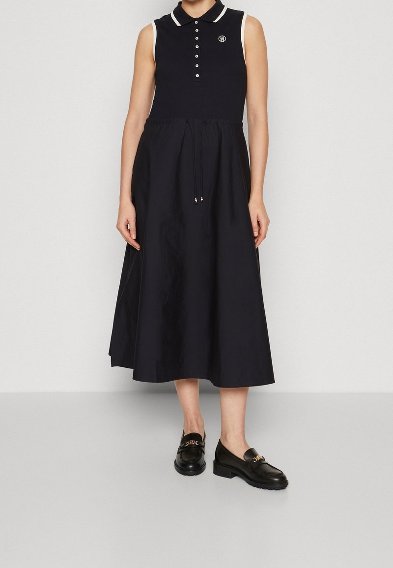 Bezramienna czarna sukienka midi z guzikiem przy kołnierzyku polo i paskiem w talii, zestawiona z czarnymi skórzanymi loafersami, na stojącej osobie.