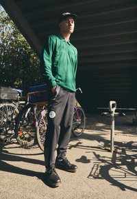 Polo de manga larga verde, pantalones negros con un parche y zapatos negros. Bicicletas al fondo; la luz del sol crea sombras en el suelo.