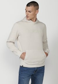 Felpa con cappuccio beige chiaro, dotata di logo testurizzato, tasca a marsupio, polsini e bordo a coste; il tessuto appare morbido e leggermente elasticizzato.