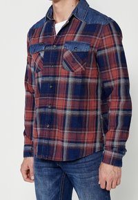 Homme portant une chemise à carreaux rouge, bleue et grise à boutons, avec col et poches en denim, associée à un jean bleu.