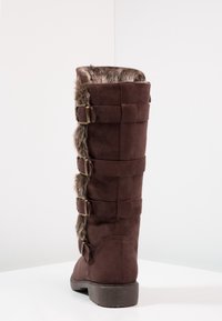Bota alta de ante marrón con borde de pelo y cuatro correas ajustables con hebillas en la parte trasera, sobre un fondo blanco liso.