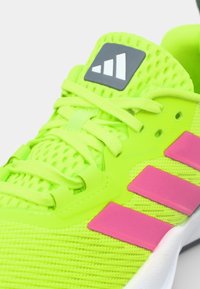 Neon grøn atletik sko med mesh tekstur, lyserøde detaljer, tre striber og en grå logo på tungen. Snørebåndene er lys grønne.