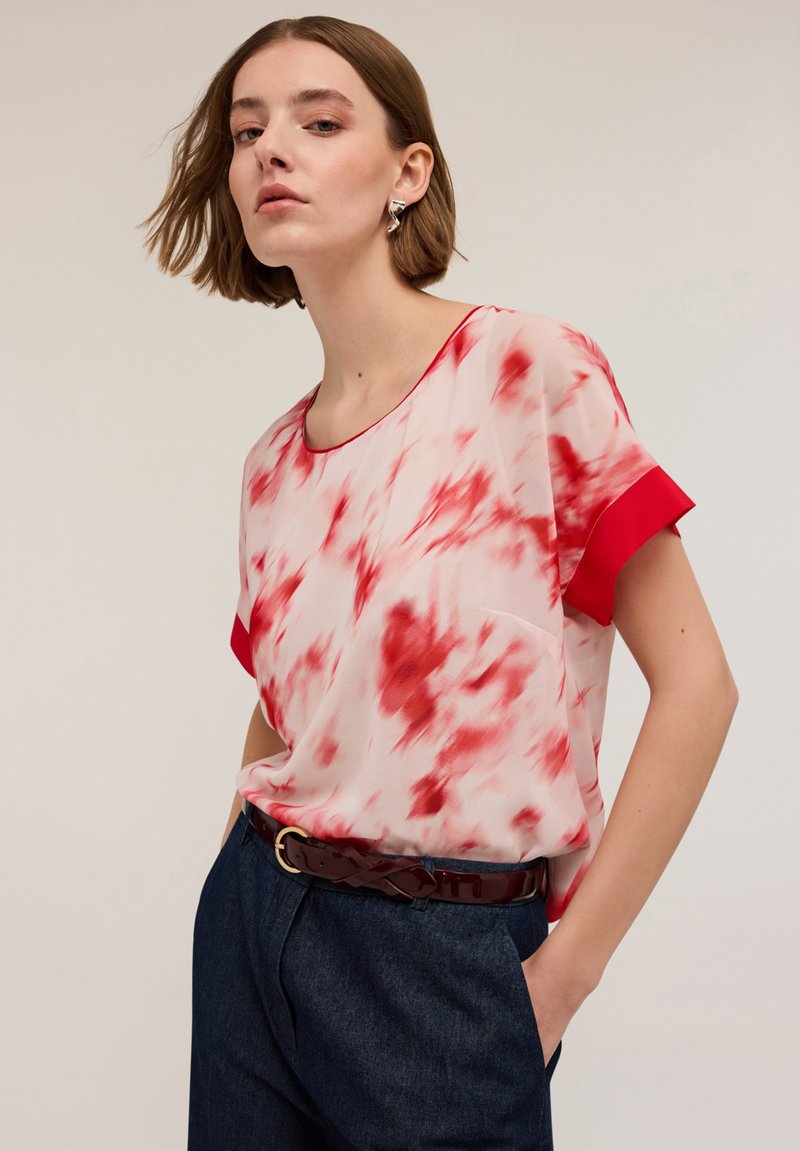 Blusa leggera in tessuto trasparente con un motivo tie-dye rosso e rosa, scollatura rotonda e maniche corte rosse. Indossata con pantaloni in denim scuri.