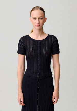 Jeune femme aux cheveux blonds tirés en arrière, portant un haut en maille noir à manches courtes boutonné et une jupe assortie sur un fond uni.