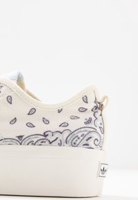 Canvas-sneakers med en krämfärgad bas, blå paisleymönster, vit sula och en liten svart och vit logotyp på hälen.
