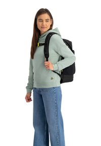 Zwarte rugzak met verstelbare banden, gekenmerkt door een gele gesp, gedragen over een lichtgroene hoodie en blauwe spijkerbroek op een witte achtergrond.