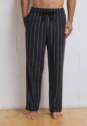 Man i svarta pyjamasbyxor med vita vertikala ränder står barfota på trägolv mot en vit tegelvägg.