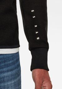 Svart texturerad tröja med ribbade ärmslut, med vit text "G S R A W" längs ärmen och en kontrasterande fåll.