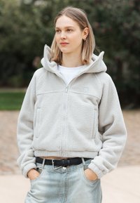 Młoda kobieta z krótkimi blond włosami, ubrana w jasnoszary polarowy rozpinany dres i paski w czarne paski, z rękami w kieszeniach, na świeżym powietrzu.
