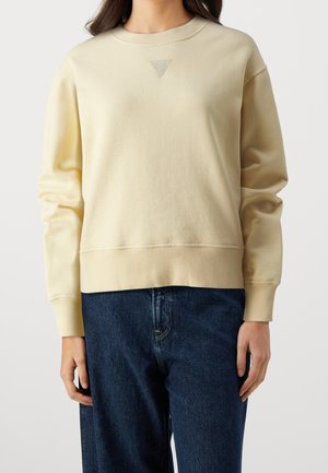 Person iført en lysebeige crewneck sweatshirt med en lille trekantsdetalje under kraven, kombineret med mørkeblå jeans.