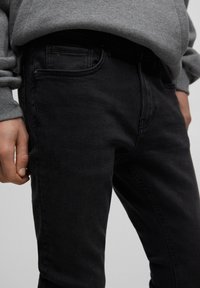 Jeans de mezclilla negros con un corte ajustado, bolsillos estándar y una textura suave. Llevados con un sudadera gris a la altura de la cintura.
