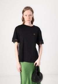 Juoda trumparegė T-shirt su sudėtingu juodu nėriniu ant rankovių, su mažuoju auksiniu logotipu ant krūtinės. Derinama su žalia sijonu.