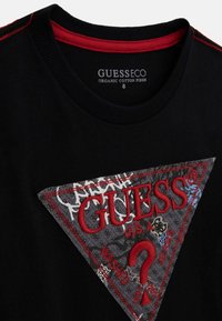 Zwarte organische katoenen t-shirt met een opvallende textuurdriehoeksgrafiek in grijs met rode "GUESS" branding en gemengde patronen. Rood accent bij de halslijn.