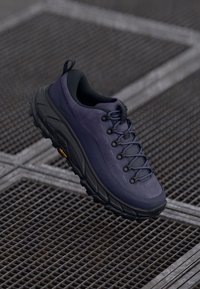 Zapatilla de senderismo de color púrpura oscuro con una parte superior texturizada, suela de goma negra y un robusto sistema de cordones. Presenta un logo amarillo en la suela.