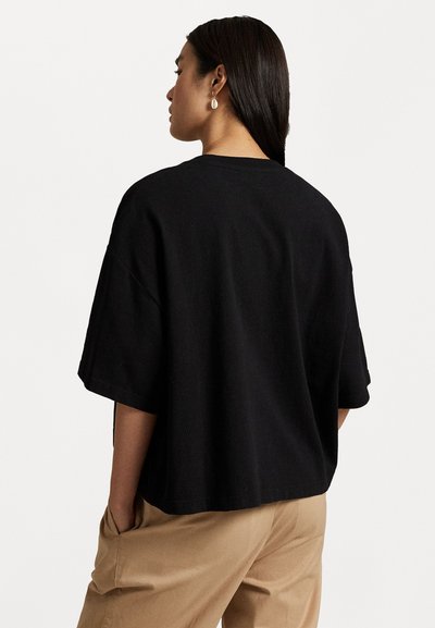 Črna oversized bombažna majica s krožnim izrezom in kratkimi rokavi, ki ima sproščen kroj in enotno teksturo. Nosijo se s bež hlačami.