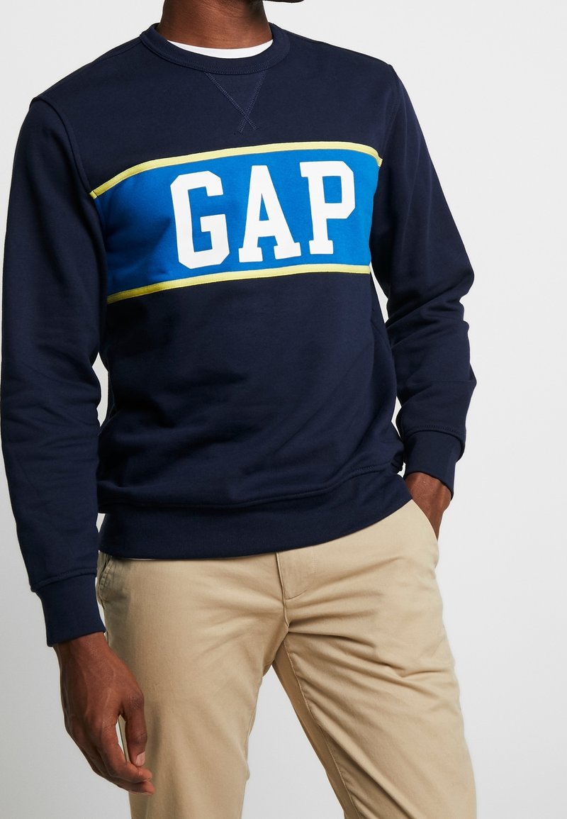 Personne à la peau foncée portant un sweat GAP bleu marine avec une bande bleue et jaune, pantalon beige, debout avec une main dans la poche sur fond blanc.