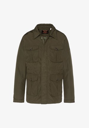 Veston vert olive avec quatre poches à rabat à boutons sur le devant, porté sur un haut noir, associé à un pantalon beige clair.
